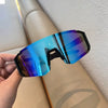 A hand holds a pair of blue-tinted, oversized sunglasses with purple and cyan reflections against a light background.