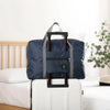 Une sac de voyage pliable bleu marine avec des sangles grises est posé sur une valise blanche à côté d'un lit.