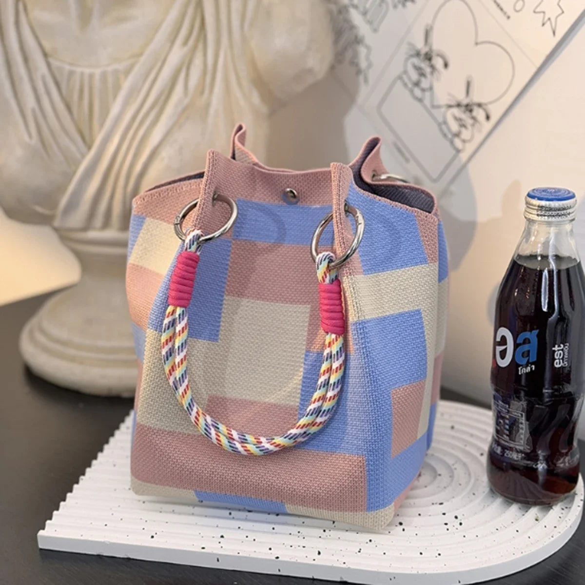 A pink and blue striped knitted bucket handbag with a woven strap resting on a white surface.