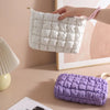 A white quilted puffer makeup bag is being held by a hand against a light pink background.
