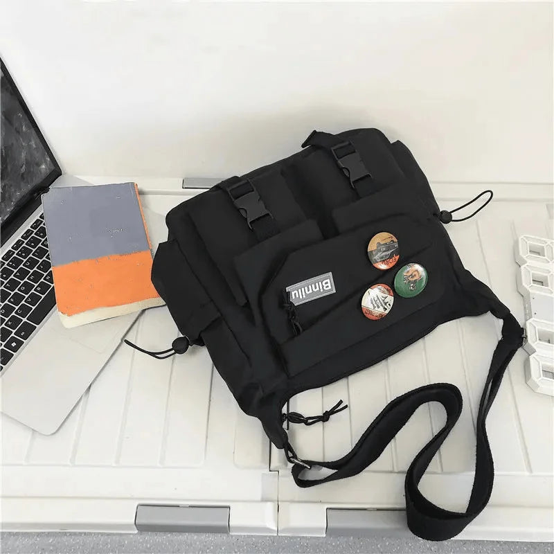 Una bolsa cruz de nylon casual negra se muestra sobre una superficie blanca junto a una computadora portátil y un cuaderno.
