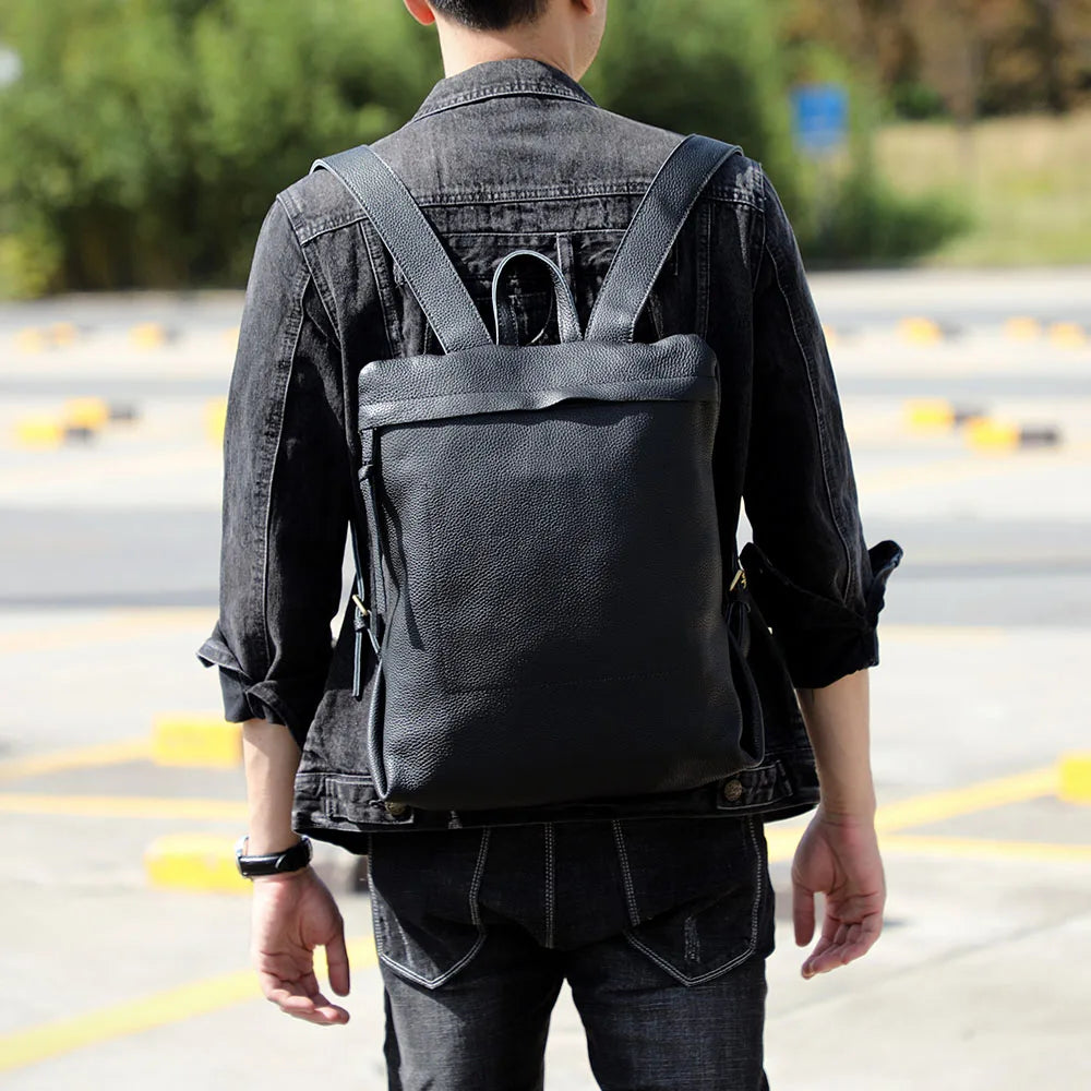A person wearing a black denim jacket and dark pants is shown from the back carrying a black full-grain leather backpack.