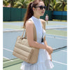 Une femme en robe blanche et lunettes de soleil se tient sur un court de tennis tenant un sac à main matelassé beige.