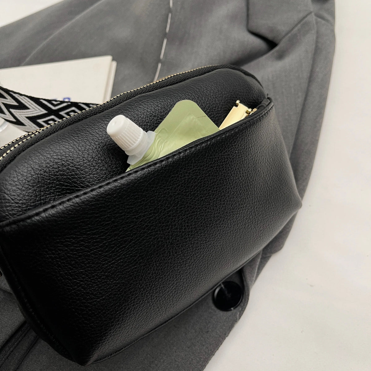 A black vegan PU crossbody bag is shown open, revealing a small white bottle and a gold item inside.