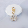 A gold-toned pendant featuring a white resin flower charm attached to a keyring.