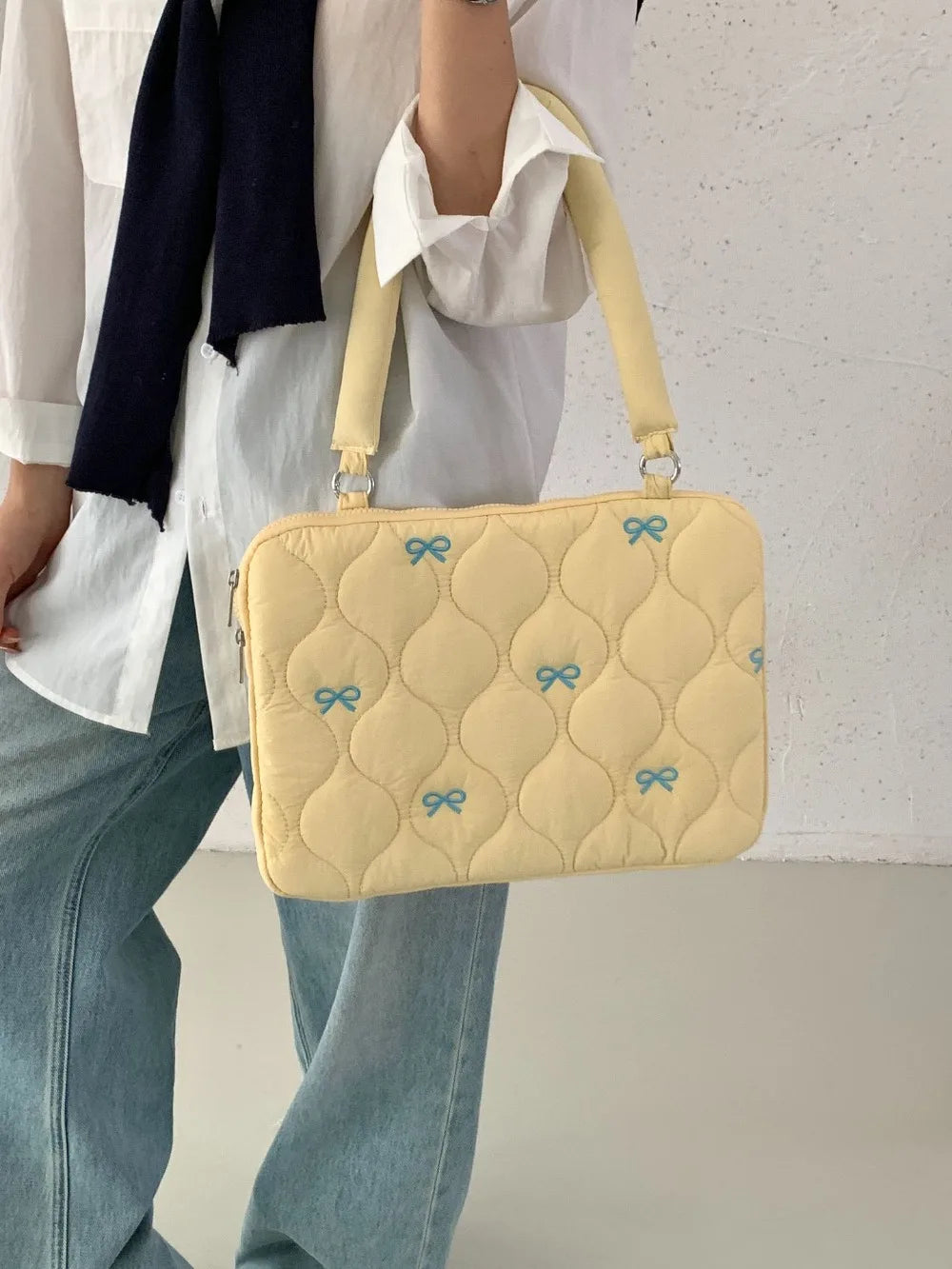 Bolso para portátil acolchado amarillo claro con detalles de lazo azul, llevado por una persona con camisa blanca y jeans.
