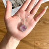 A hand holds a handcrafted amethyst crystal pendant necklace featuring a pinkish-purple gemstone set in a silver-toned wrap.