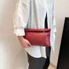 Una mujer sostiene un bolso de mano rectangular tejido en color rojo intenso contra una camisa blanca y pantalones oscuros.