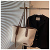 A beige canvas shoulder tote bag with brown leather straps hanging on a hanger against a window blind.