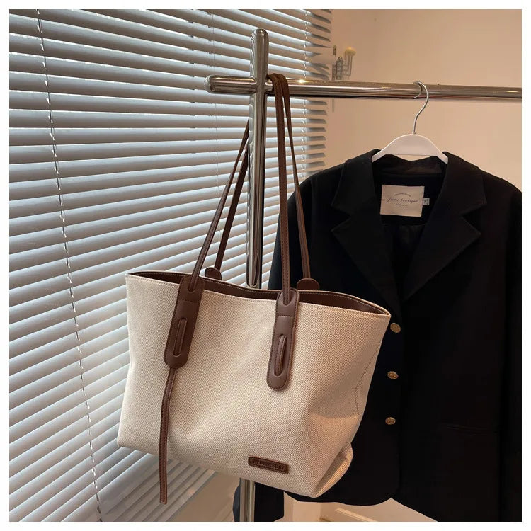 A beige canvas shoulder tote bag with brown leather straps hanging on a hanger against a window blind.