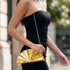 A woman wearing a black dress holds a gold acrylic chain crossbody bag with a fan-like design.