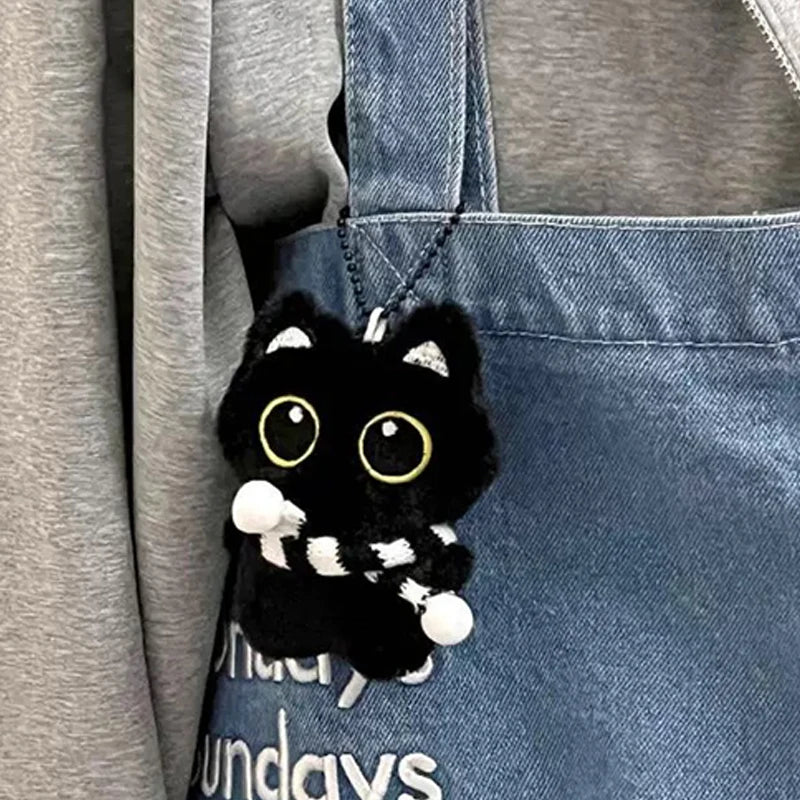 Colgante de gata negra de peluche con detalles blancos, colgando de una bolsa de tela vaquera.