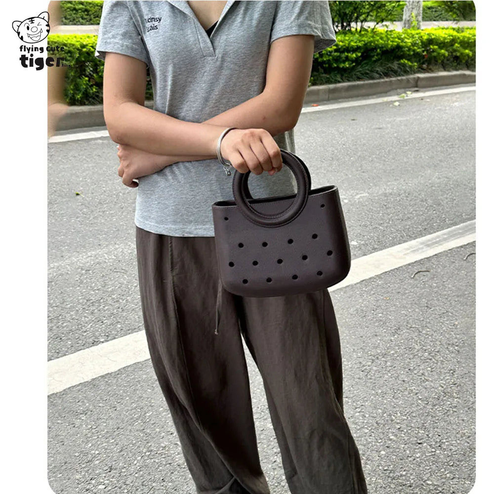 A woman is holding a dark brown round PVC handbag with a DIY hole design while standing outdoors.