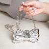 A hand is holding a silver acrylic bow clasp mini handbag with a chain strap on a light surface.
