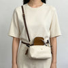 A woman wearing a cream-colored top and trousers, carrying a brown leather crossbody bag with a detachable mini pouch.