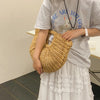 A person holds a woven rattan half-moon beach handbag against a light gray t-shirt and white skirt.