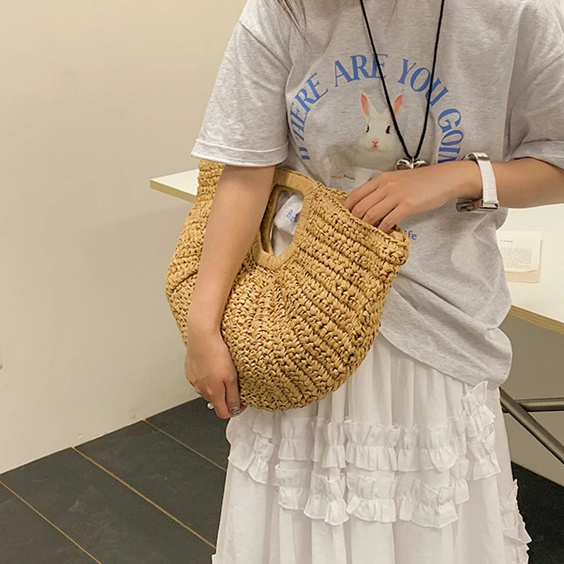 A person holds a woven rattan half-moon beach handbag against a light gray t-shirt and white skirt.