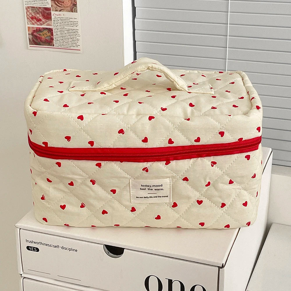 A cream quilted makeup bag with a red trim and small red hearts, resting on a white box.
