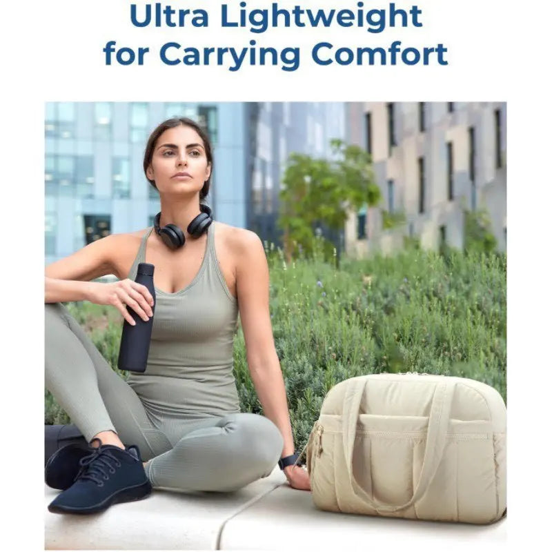 A woman is sitting outdoors wearing a olive green top and light pants, holding a beige travel bag.