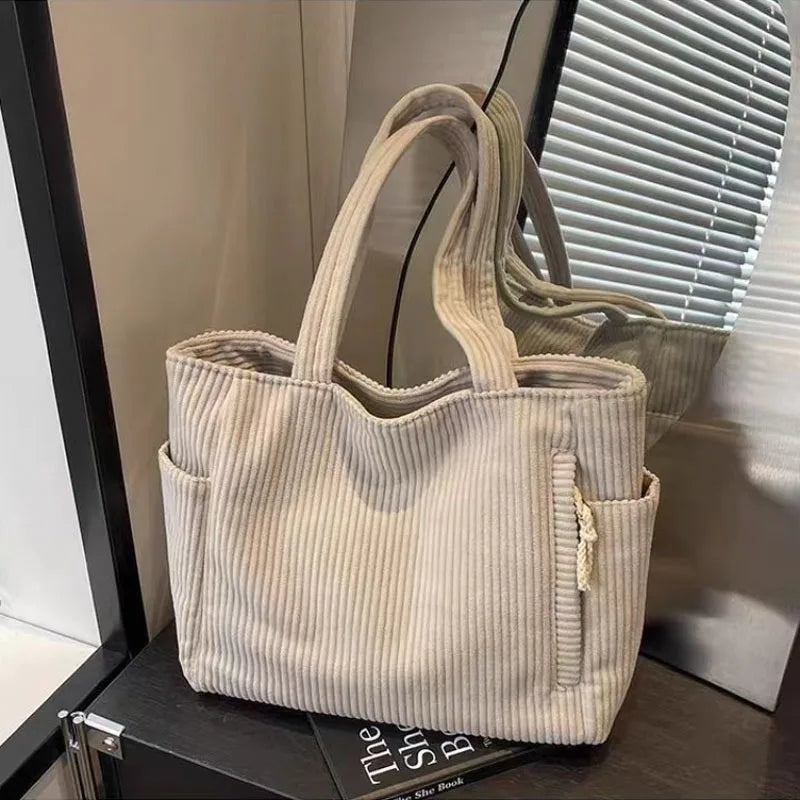 A beige corduroy shoulder bag with a zipper is displayed on a dark surface next to a window.
