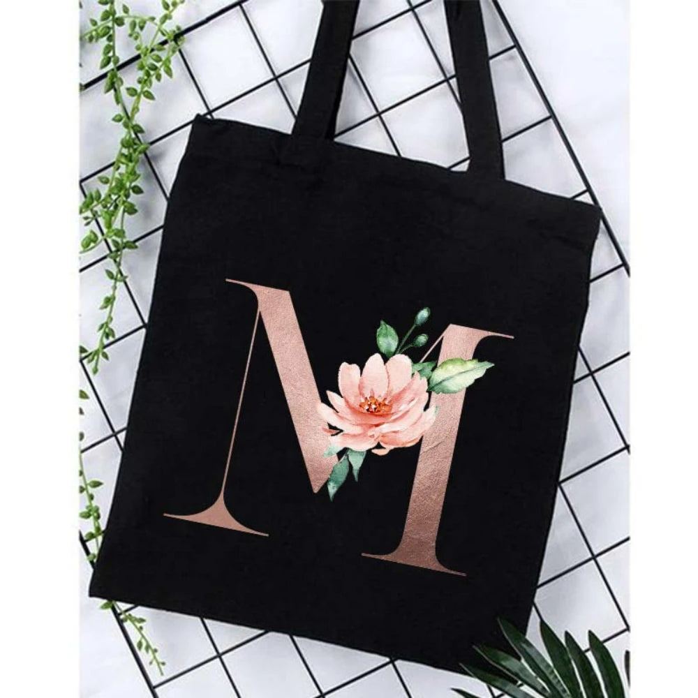 Black canvas tote bag featuring a large gold letter M with pink floral accents