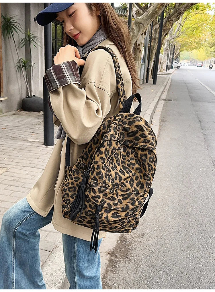 A woman wearing a beige jacket and jeans is carrying a leopard print canvas backpack while walking on a sidewalk.