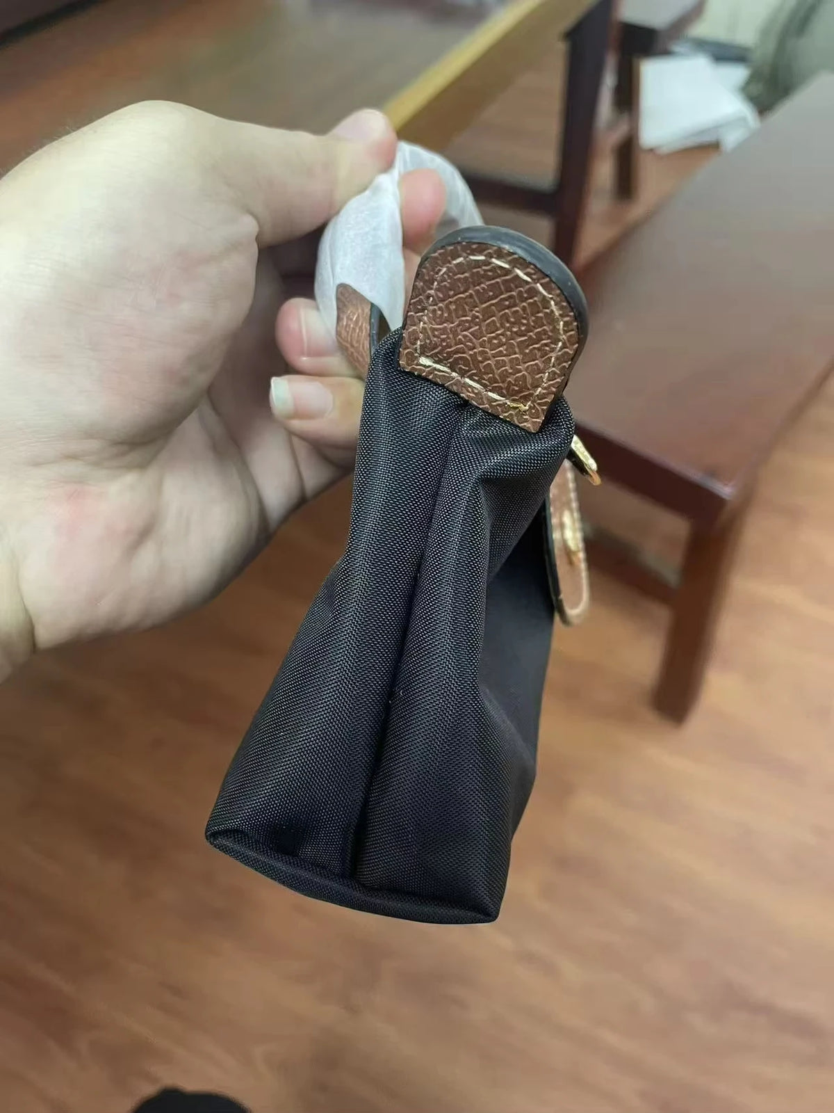 A hand holds a black nylon crossbody bag with a brown leather detail and a white strap.