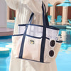 A white mesh beach tote bag with navy trim is held by a person near a swimming pool.