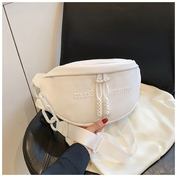A white PU leather crossbody saddle bag with a chain strap resting on a black leather surface.