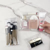 A transparent portable organizer pouch holds makeup brushes, a perfume bottle, and a cosmetic product.