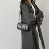 A woman wearing a gray coat holds an elegant black leather tote bag with the word KURT embossed on it.