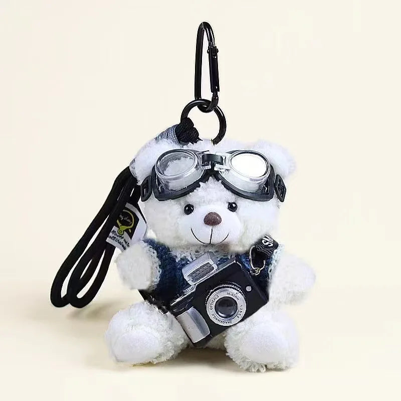 A white plush bear keychain wearing black goggles and holding a small camera.