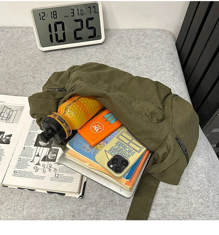 A olive green cotton canvas shoulder crossbody bag is shown open on a gray surface next to a digital clock and some papers.