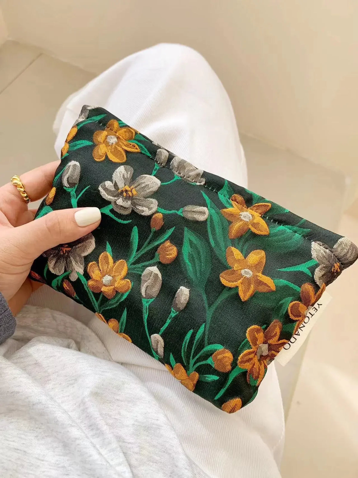 A dark green cosmetic pouch featuring a floral pattern of yellow and gray flowers being held by a hand.