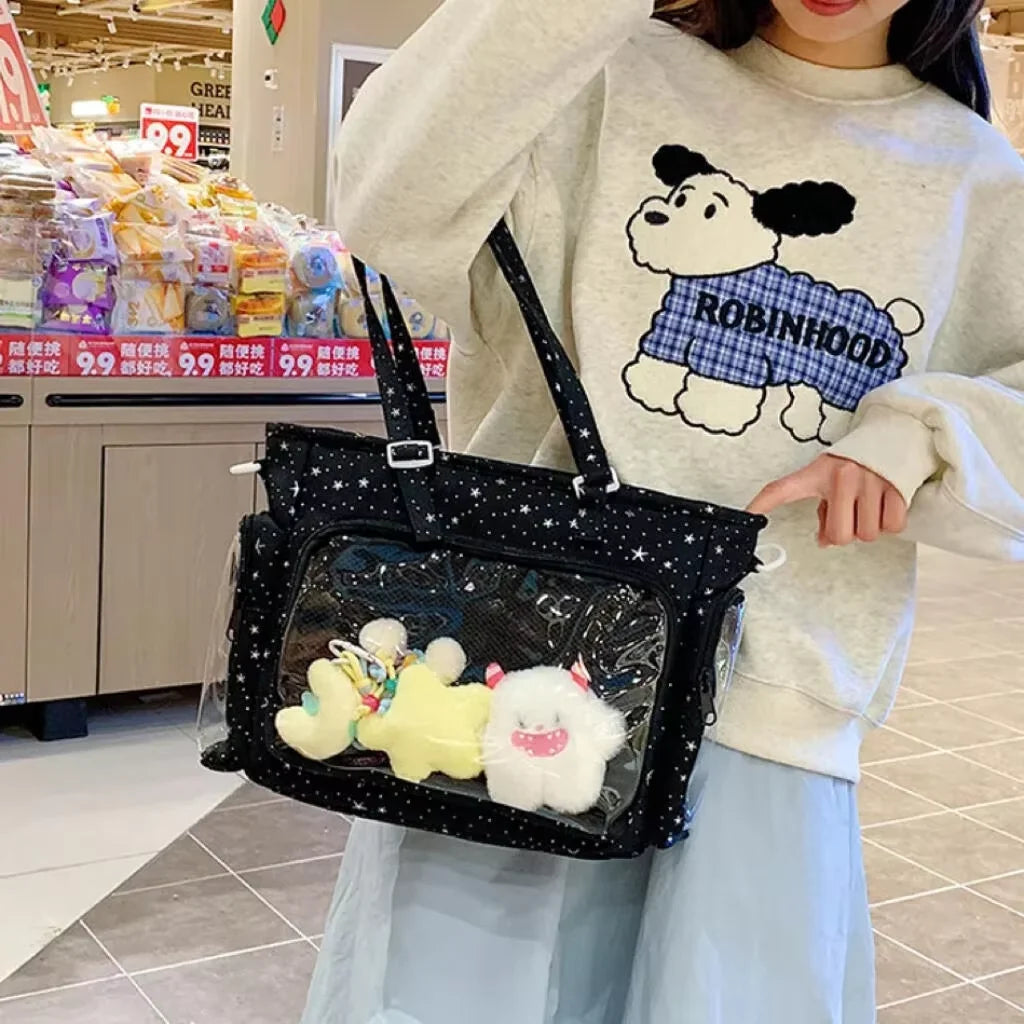 A person holds a black transparent shoulder tote bag with a white fluffy animal charm against a blurred background.