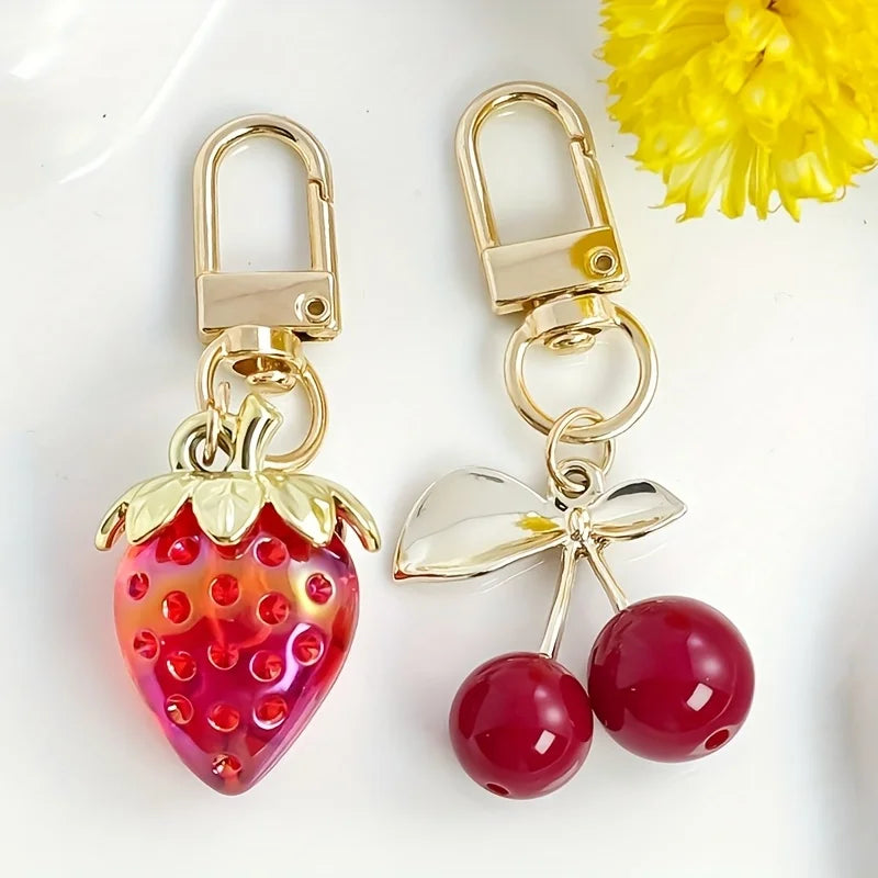 A set of gold-toned keychain charms featuring a strawberry and a cherry, with a yellow flower in the background.
