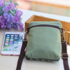 A green denim mini messenger bag with brown leather straps resting next to a smartphone and purple flowers.