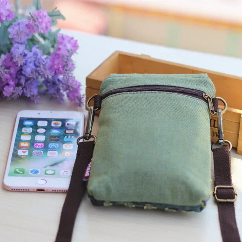 A green denim mini messenger bag with brown leather straps resting next to a smartphone and purple flowers.