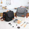 A gray insulated reusable lunch bag with brown straps rests on a white table next to cutlery and small dark fruit.