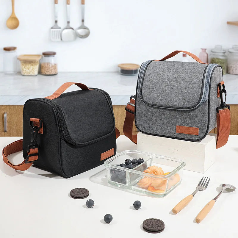 A gray insulated reusable lunch bag with brown straps rests on a white table next to cutlery and small dark fruit.