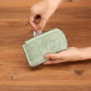 A hand is opening a light green floral patterned zip coin purse with a silver key ring on a wooden surface.