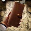 A hand holds a brown genuine leather long bi-fold wallet with a snap closure on a textured paper surface.