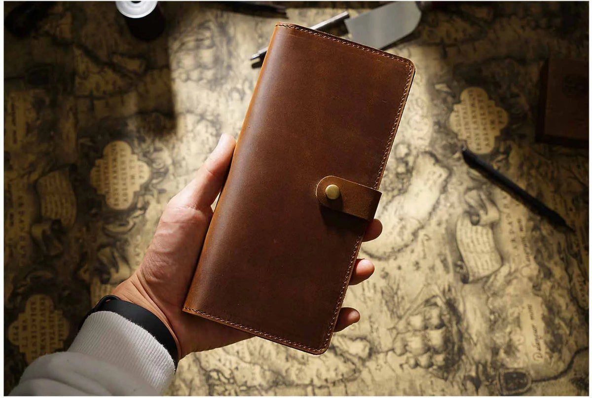 A hand holds a brown genuine leather long bi-fold wallet with a snap closure on a textured paper surface.