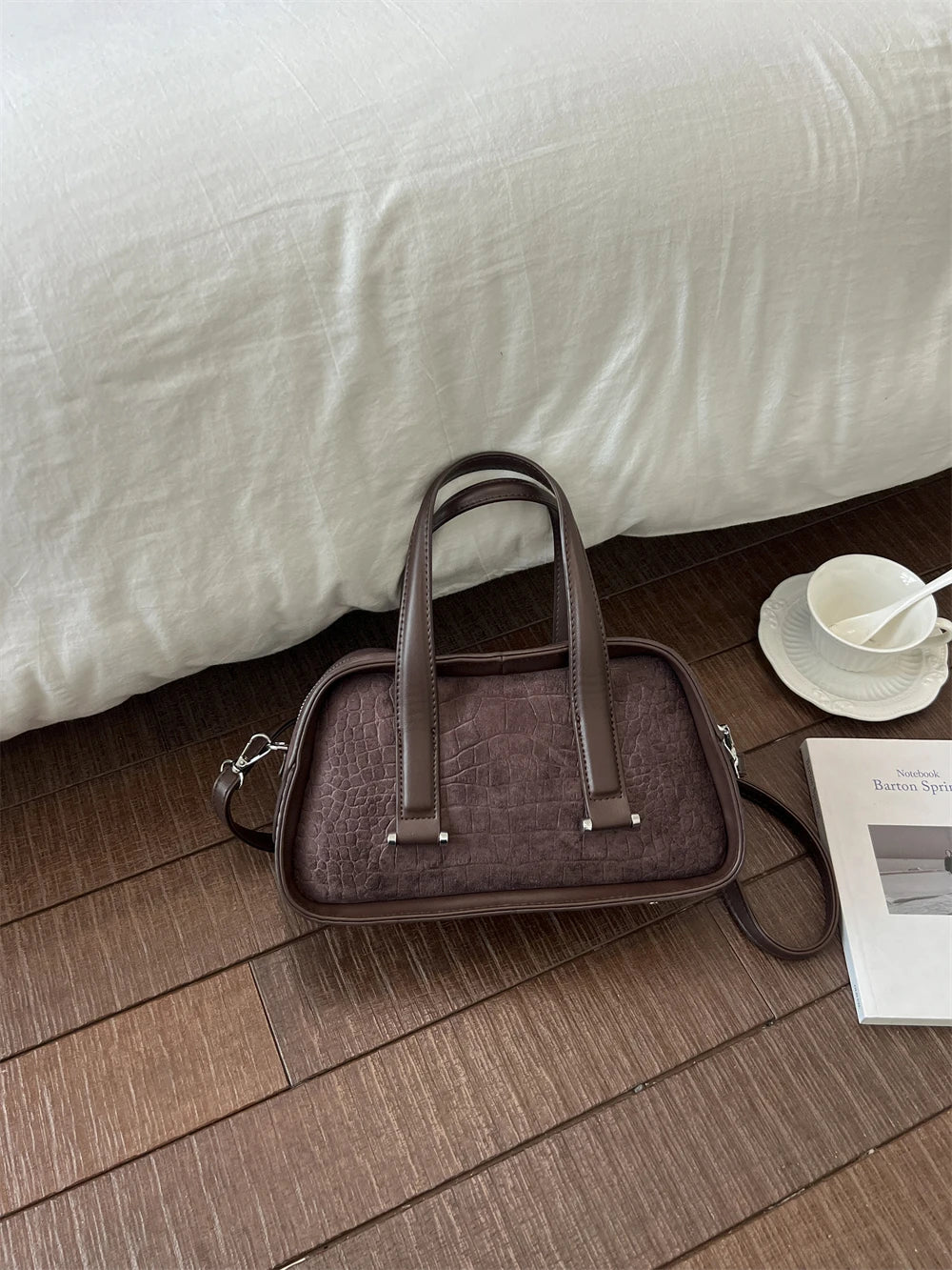 A brown leather mini crossbody bag with a flap and handles resting on a wooden floor next to a white cup.