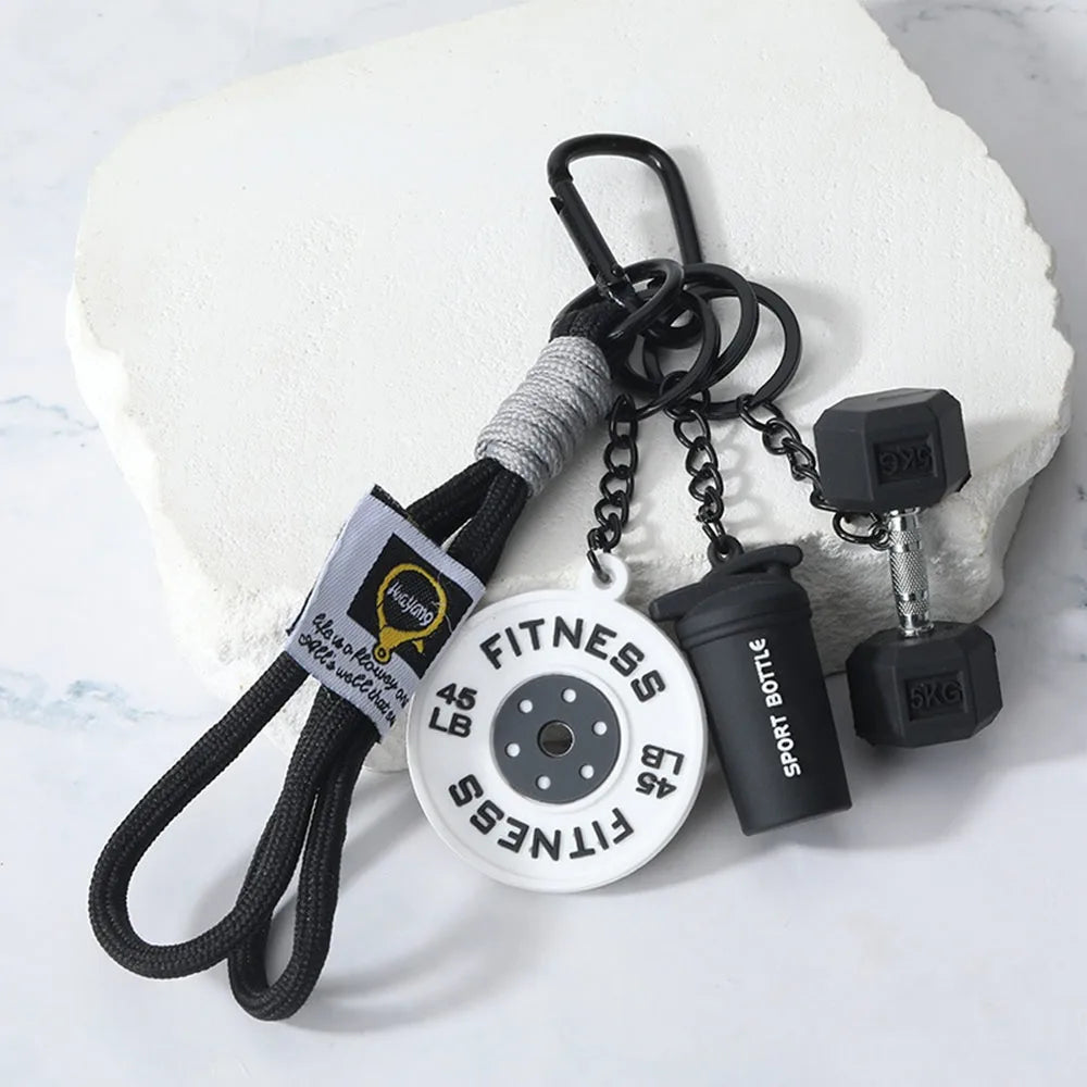 A black detachable dumbbell keychain charm with a fitness disc and a sports bottle attached to a white block.