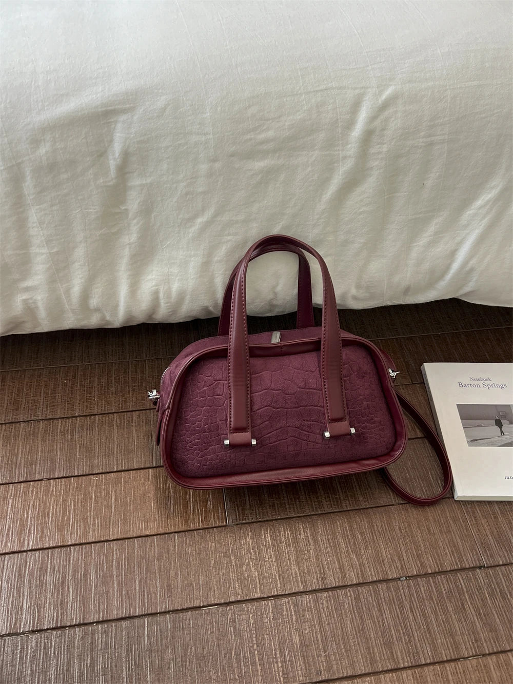 A maroon retro crocodile textured mini crossbody bag resting on a wooden floor next to a white box.