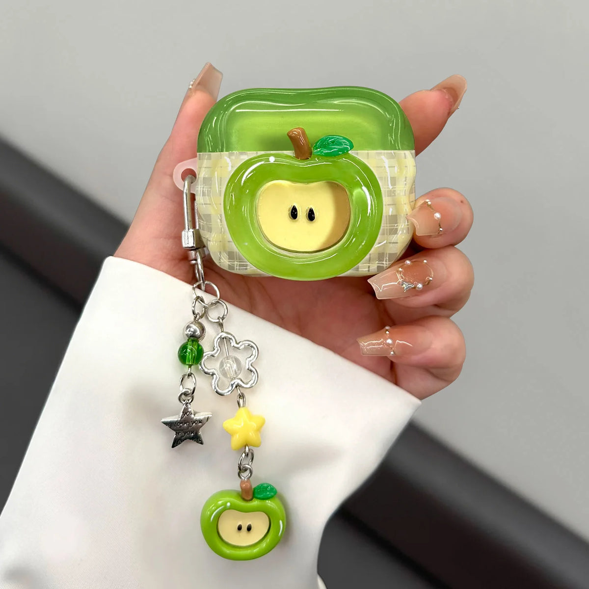 A hand holds a green apple shaped earphone case with a yellow interior and a silver charm necklace.