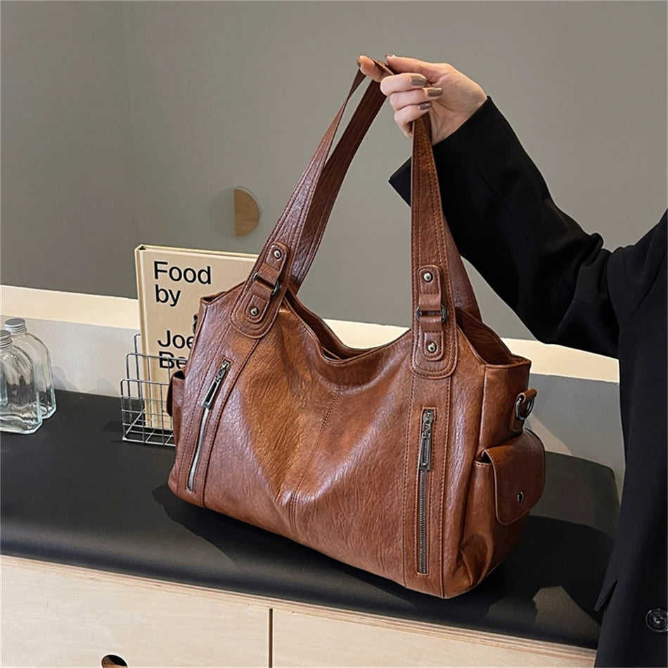 Une personne tient un sac à main en cuir marron avec plusieurs poches sur un fond clair.