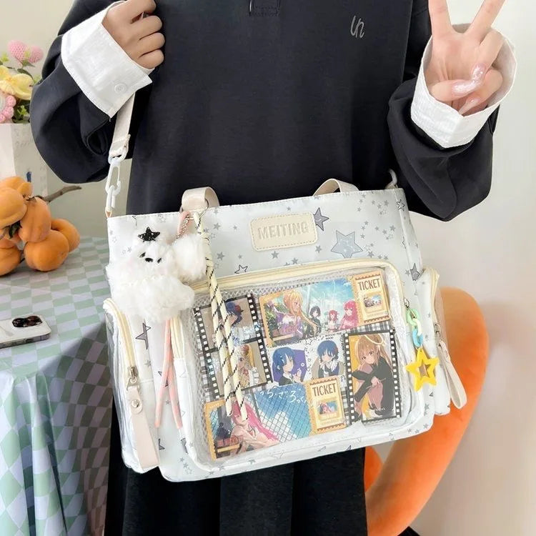 A person holds a white transparent shoulder tote bag with various ticket-themed patches and fluffy white pom-poms.