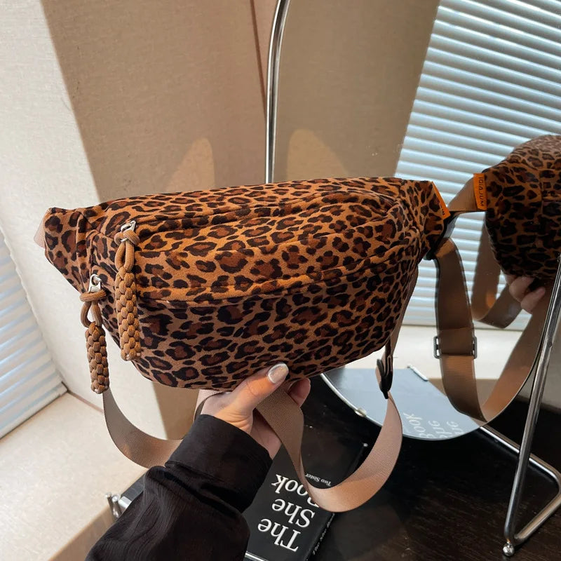A leopard print crossbody bag with a brown adjustable strap is displayed on a silver stand.